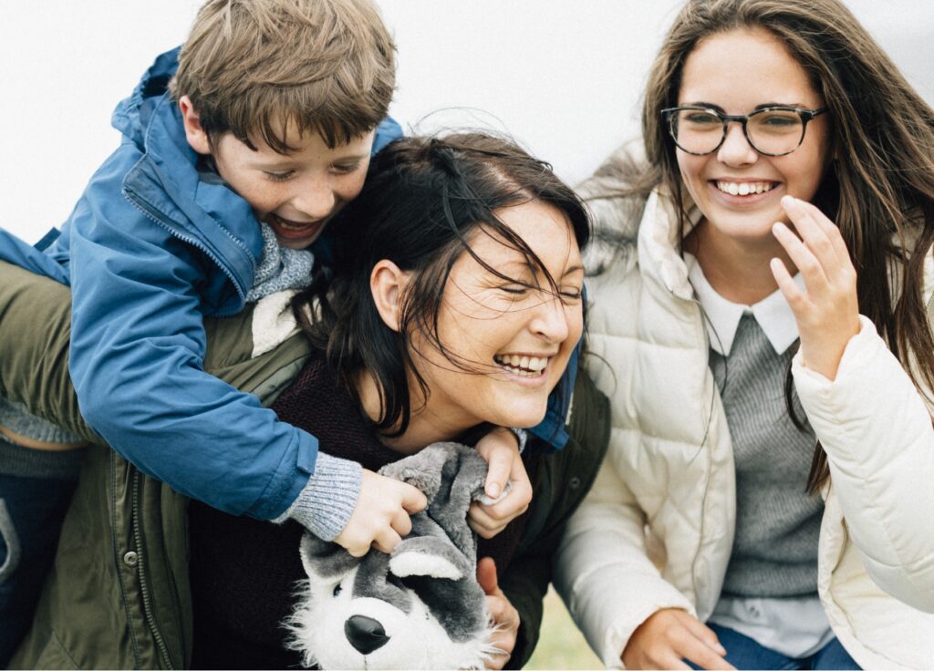 a young family playing outside together, they're happy. the small boy climbs on the mothers back, and hugs her to hold on. The daughter smiles and is wearing glasses. It is cold outside and they are warm.
